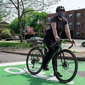 A bicyclist stopped on pavement painted green with a bike symbol. The bicyclist is wearing a face mask.